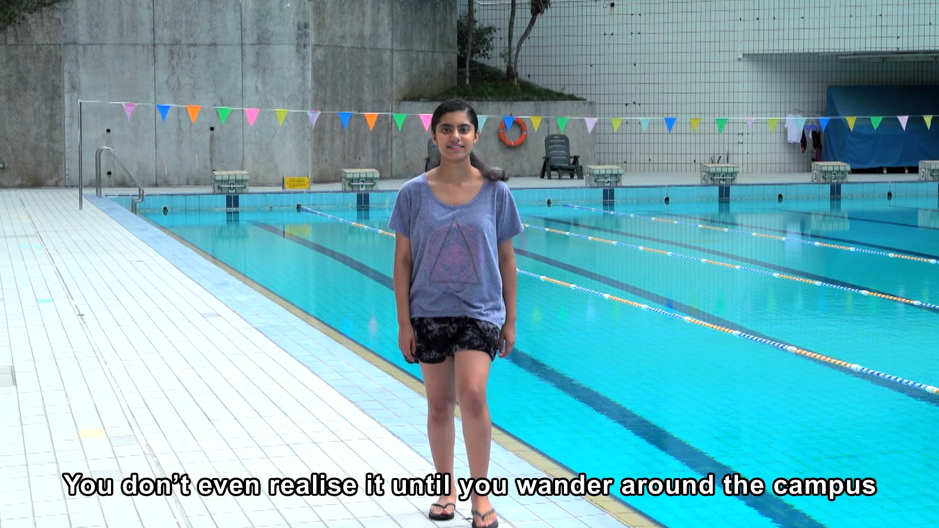 A young woman stands by an outdoor swimming pool, smiling at the camera. She wears a loose T-shirt, shorts, and sandals. Colorful bunting decorates the pool area. Subtitle: "You don’t even realise it until you wander around the campus.