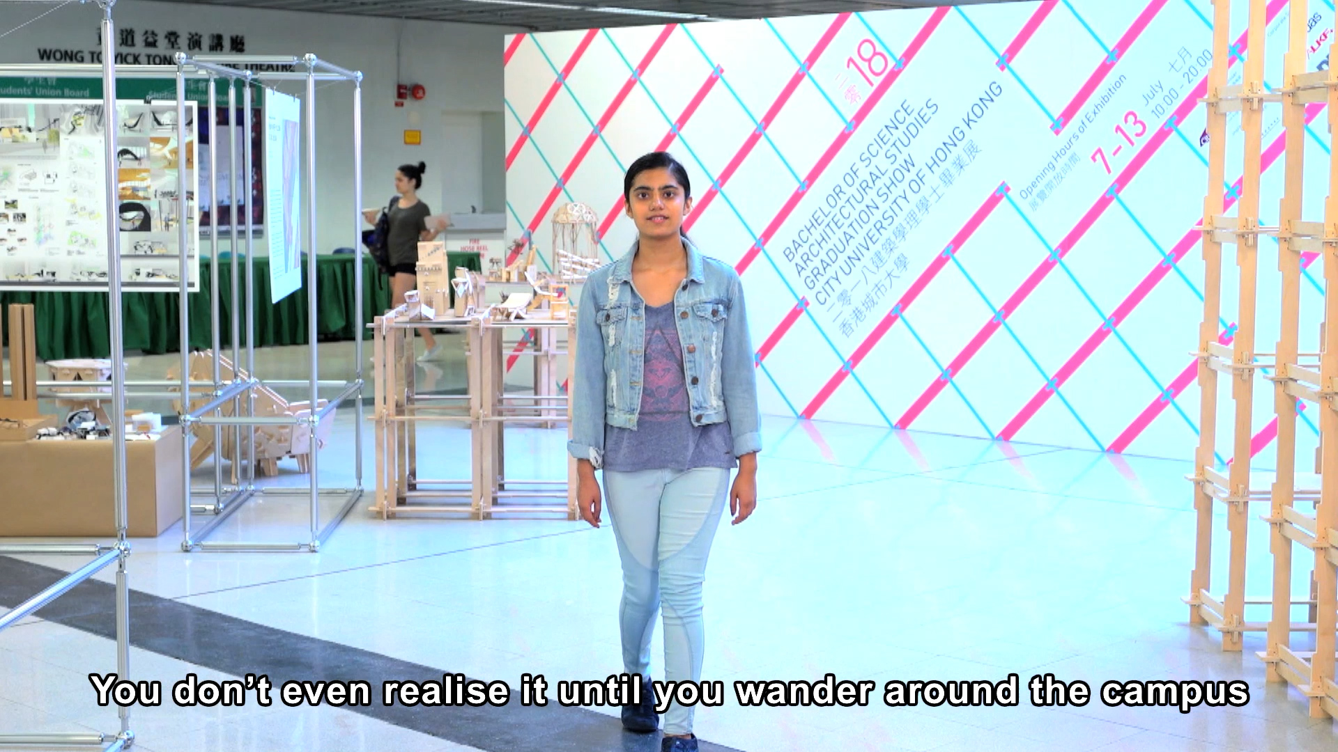 A young woman in light blue jeans and a denim jacket stands indoors at an exhibition. Architectural models are displayed around her. Subtitle reads, “You don’t even realise it until you wander around the campus.”.