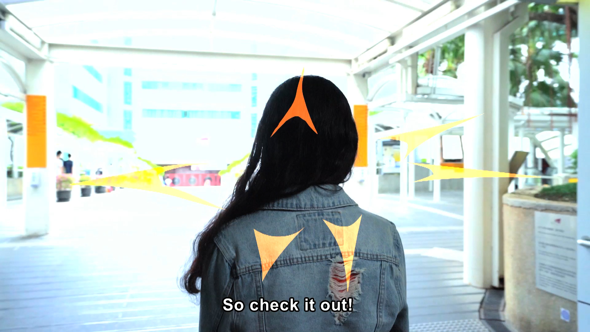 A person with long dark hair, wearing a denim jacket, stands with their back to the camera in a bright outdoor walkway. Yellow-orange graphic arrows point toward them, and text at the bottom reads, "So check it out!.