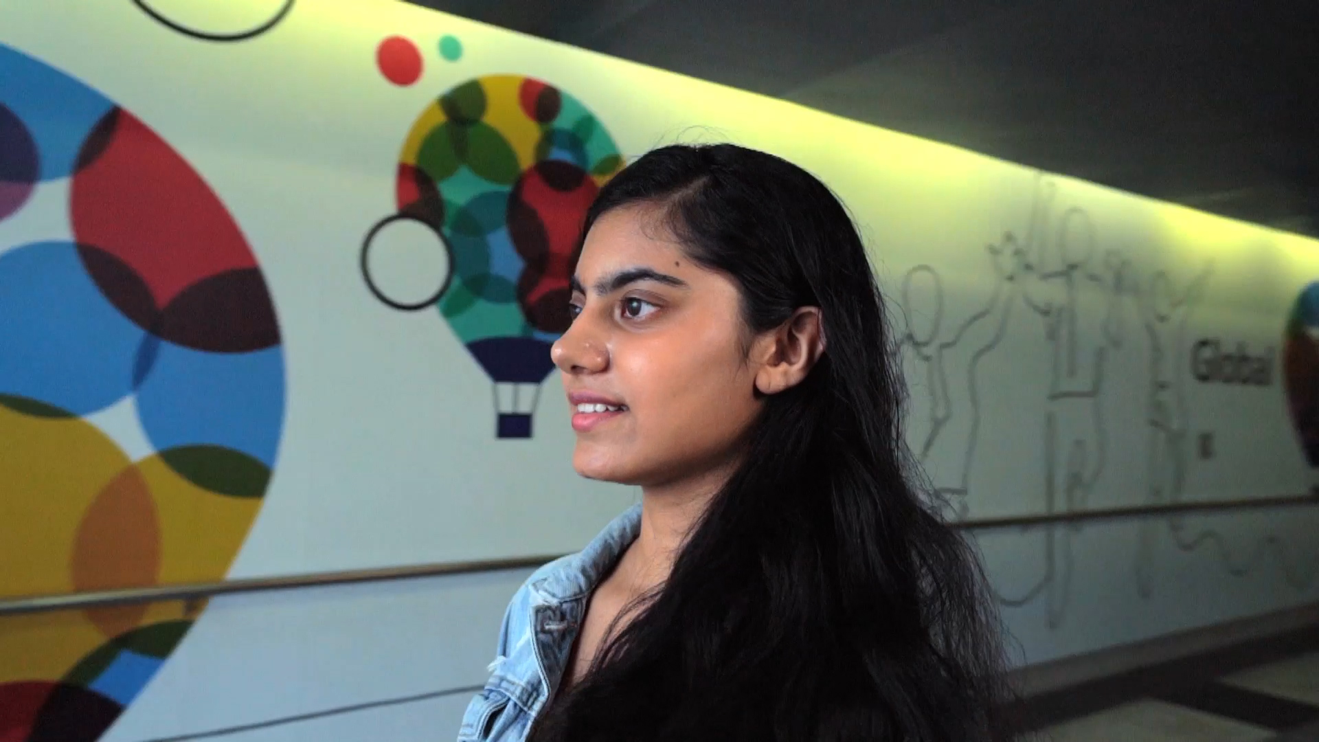 A young woman with long dark hair, wearing a denim jacket, stands indoors in front of a colorful mural featuring a hot air balloon design and abstract human outlines on the wall.