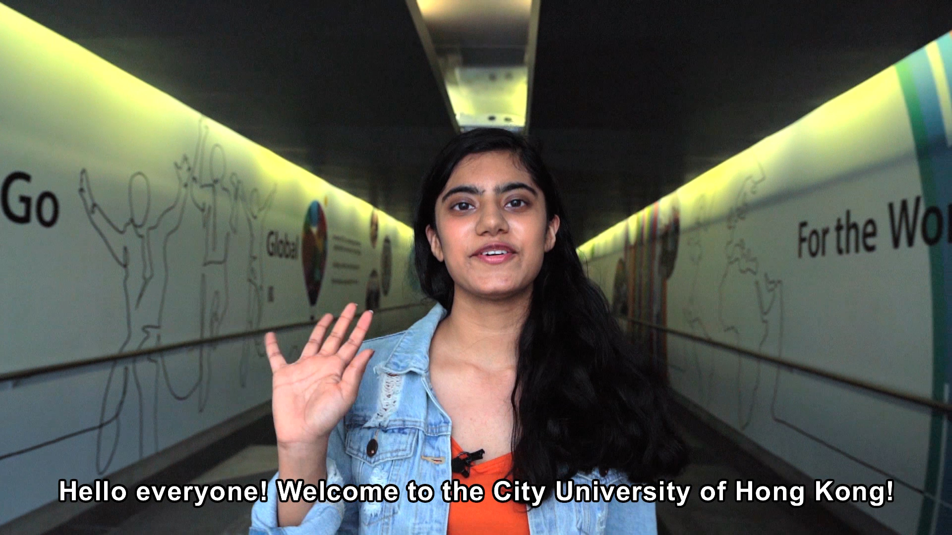 A young woman with long dark hair, wearing a denim jacket, stands in a hallway with wall art and waves at the camera. Text at the bottom reads, "Hello everyone! Welcome to the City University of Hong Kong!.