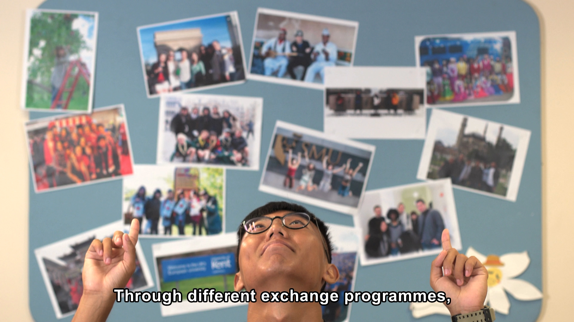 A person wearing glasses points upward with both hands, standing in front of a bulletin board covered with various group photos. Subtitle text reads: "Through different exchange programmes,.