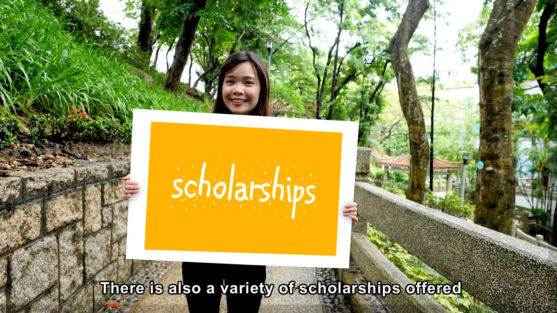 A smiling young woman stands on a tree-lined path holding a large yellow sign that reads "scholarships." Subtitle at the bottom says, "There is also a variety of scholarships offered.