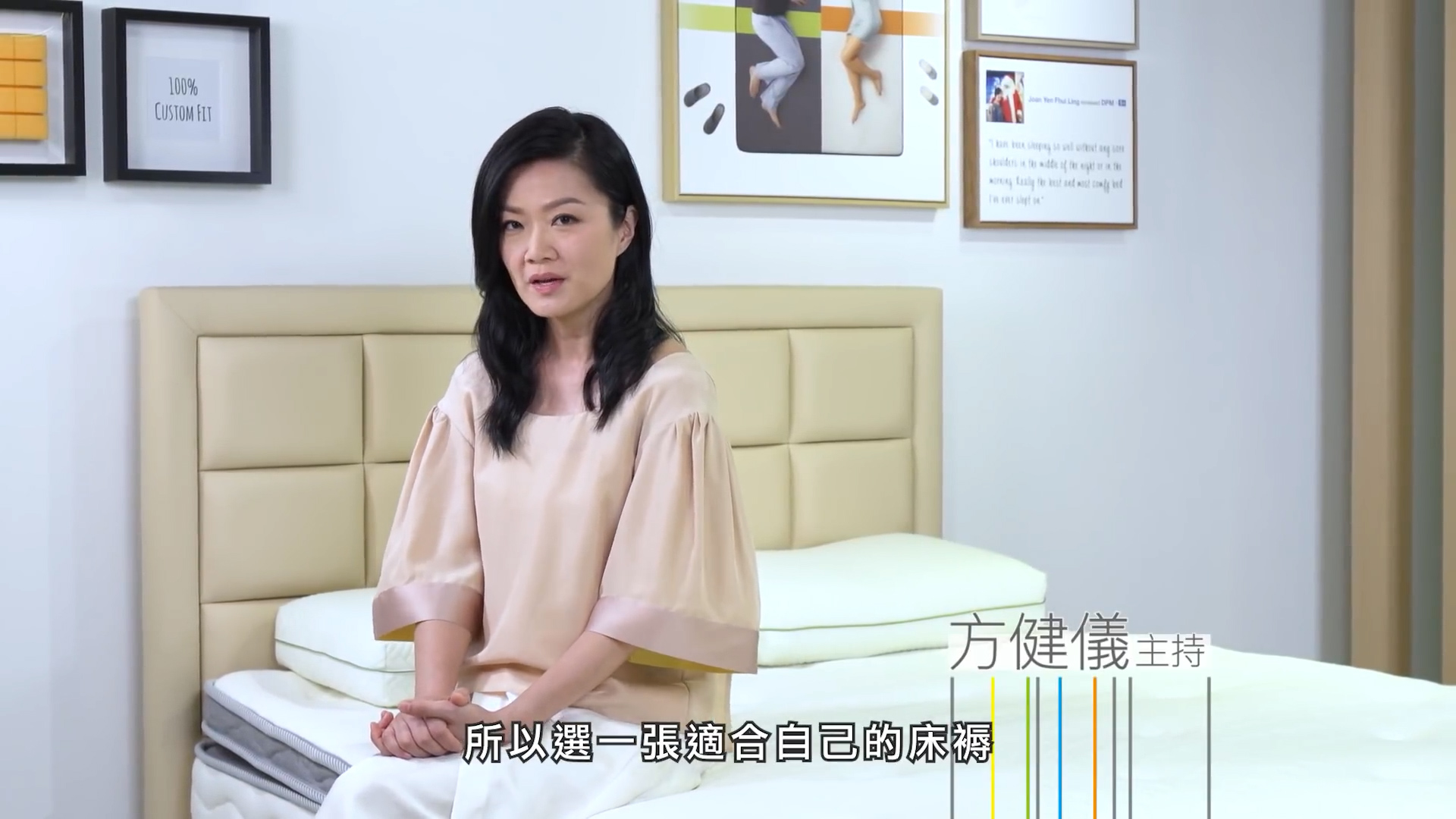 A woman with long black hair sits on a bed in a bright showroom with mattresses and framed pictures on the wall. Chinese subtitles and the name "方健儀" appear on the screen.