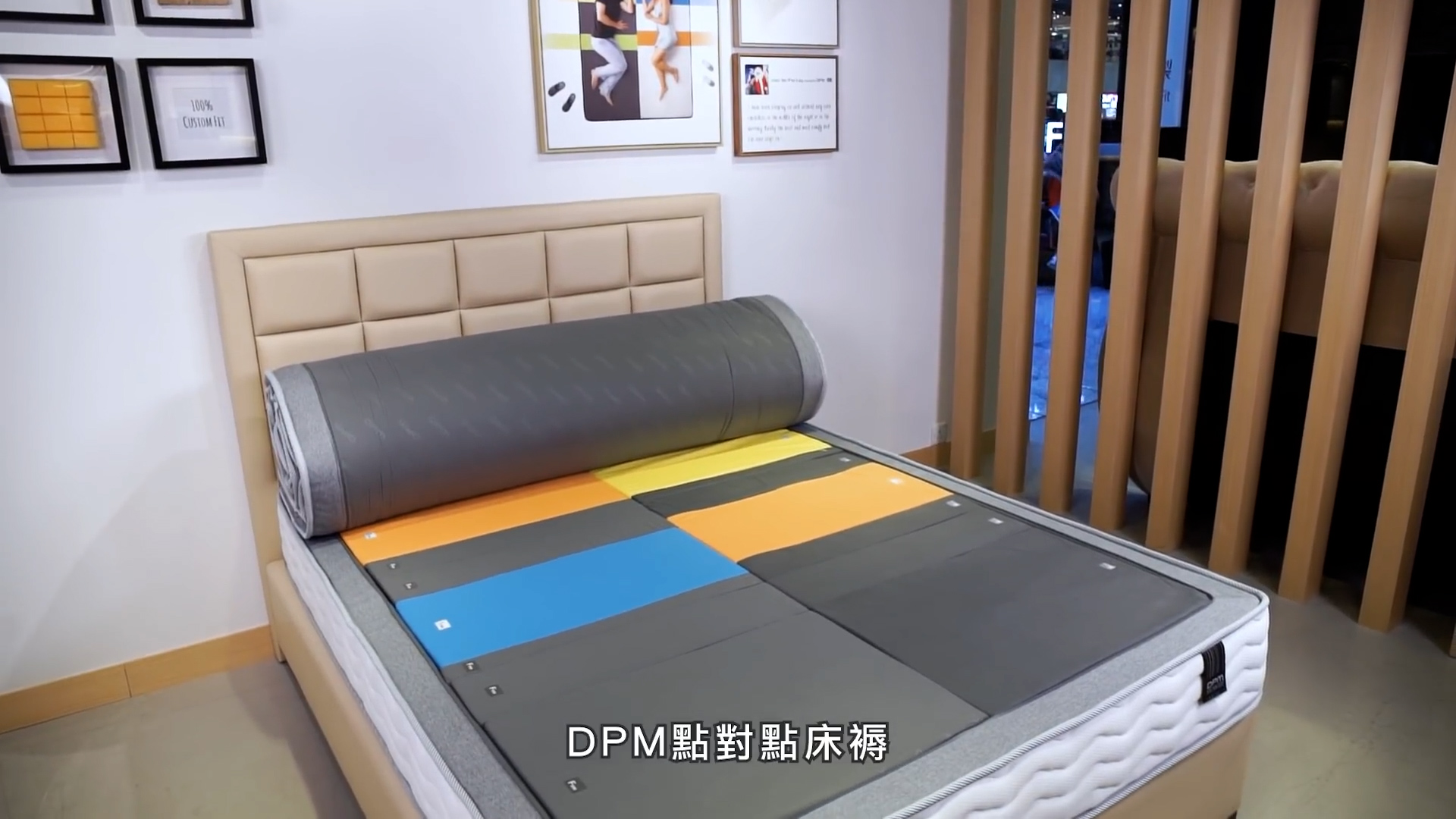 A modern bed is displayed in a showroom, featuring a colorful, segmented mattress with blue, yellow, and orange sections. Framed pictures hang on the wall, and Chinese text appears at the bottom of the image.