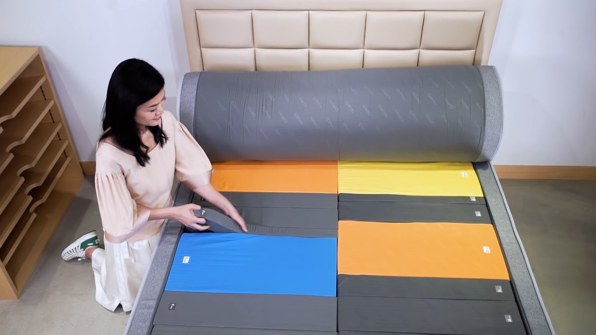 A woman arranges colorful rectangular panels—blue, orange, and yellow—on a gray cushioned surface in a bedroom, next to a beige upholstered headboard.