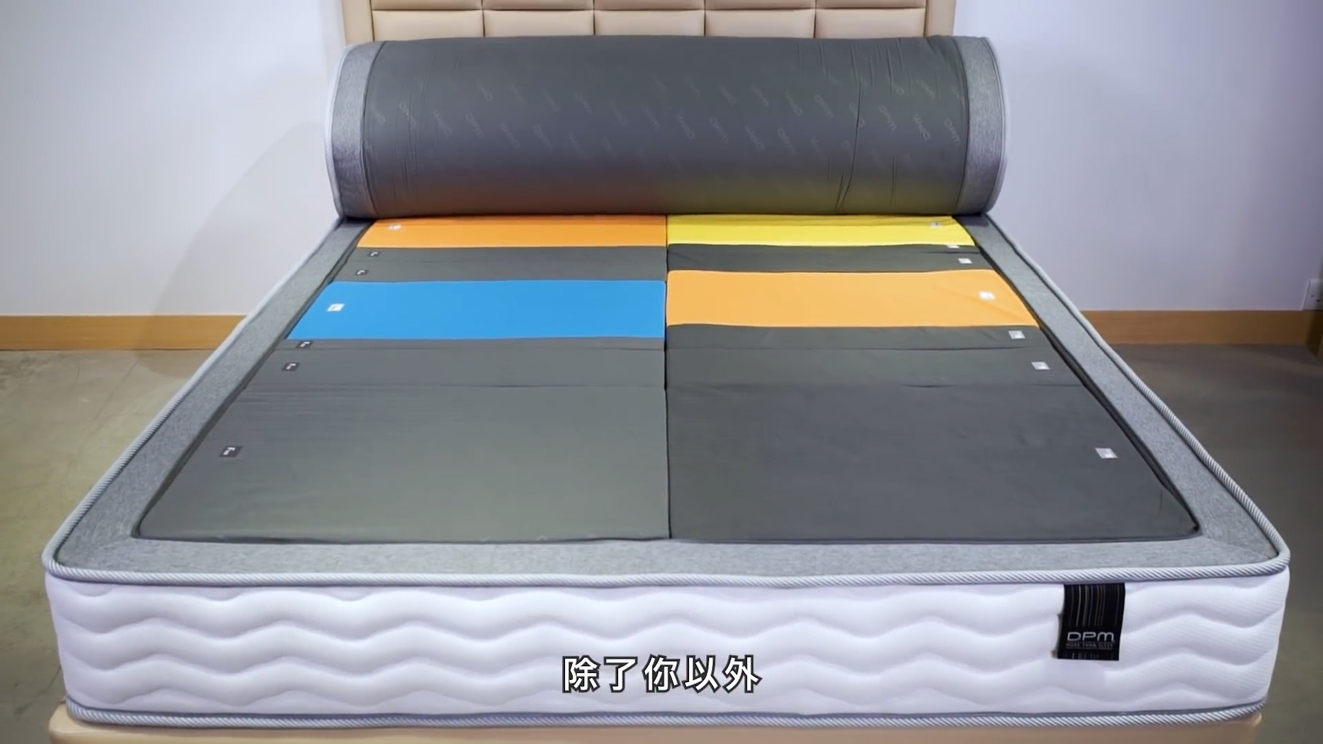A mattress with a large cylindrical pillow at the head and six colored sections—blue, yellow, orange, and black—on top. Chinese text is displayed at the bottom of the image.