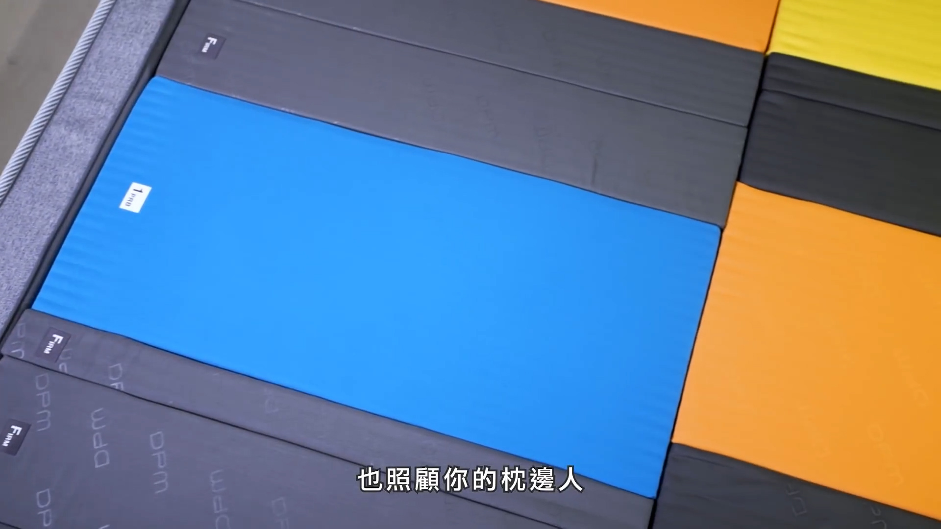 A variety of rectangular sleeping pads in blue, yellow, orange, and shades of gray are arranged side by side. Chinese subtitles are visible at the bottom of the image.