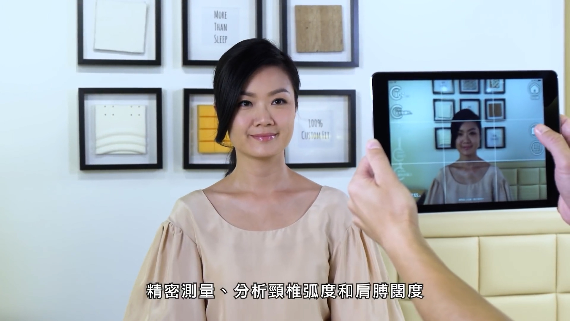 A woman in a beige dress smiles while posing for a photo taken with a tablet. Framed artwork and photos are on the wall behind her. Chinese subtitles appear at the bottom of the image.
