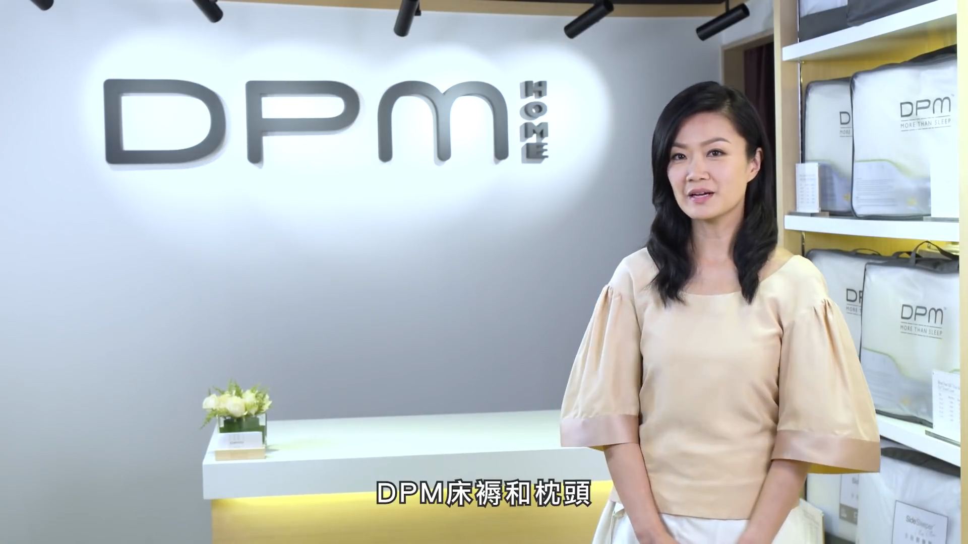A woman stands in a modern showroom with "DPM HOME" on the wall behind her. She is wearing a beige blouse and white skirt. On-screen Chinese text reads “DPM 床褥和枕頭”. Pillows and bedding are displayed on shelves to the right.