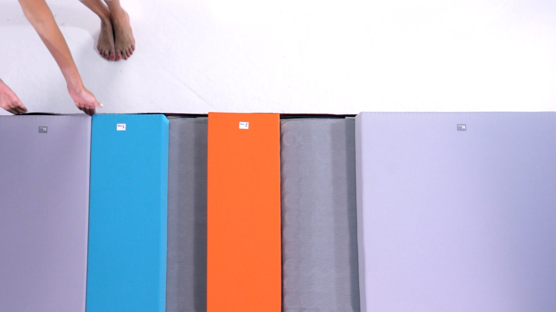 A person’s bare feet and hand are visible next to three rectangular mats—one blue, one orange, and one gray—on a white floor.