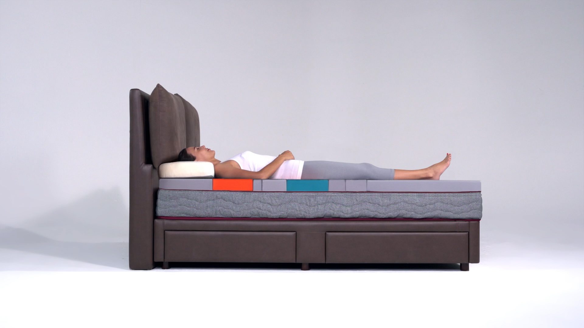 A person lies on their back on a bed with a tall headboard, demonstrating different colored sections of a mattress, likely showcasing various layers or support zones, in a minimalist studio setting.