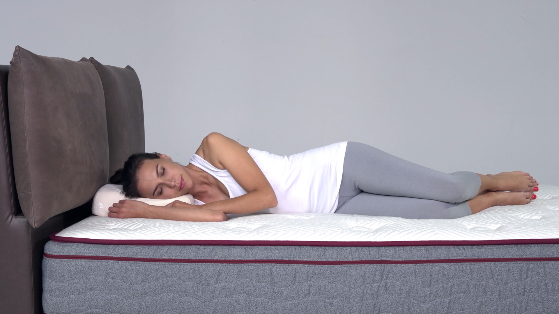 A woman wearing a white tank top and gray leggings is sleeping on her side on a bed with a white mattress and a dark headboard, resting her head on a pillow.