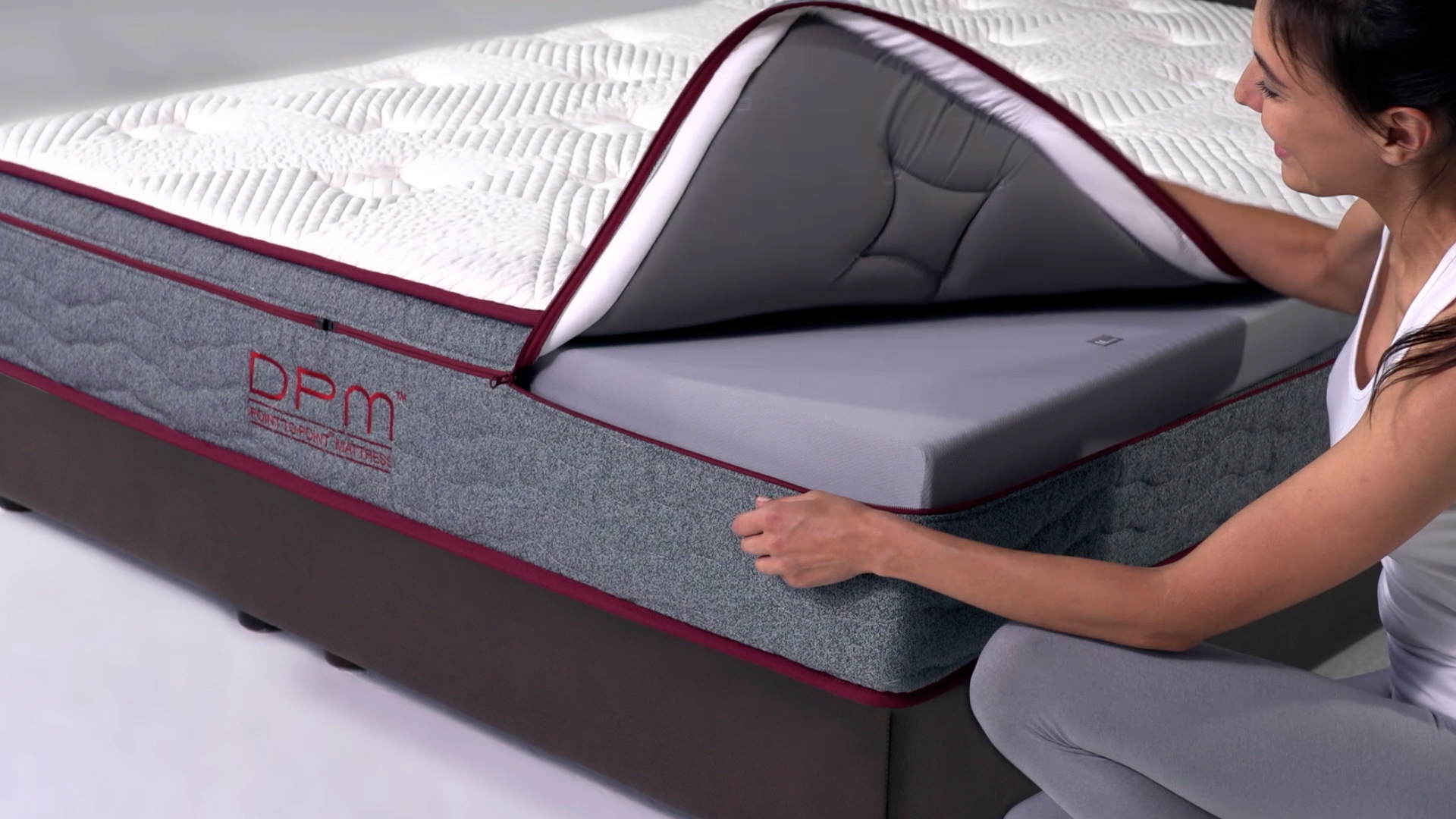 A woman in gray clothing lifts the top layer of a mattress, revealing a gray foam layer inside. The mattress features a zippered cover and is labeled "DPM" on the side.