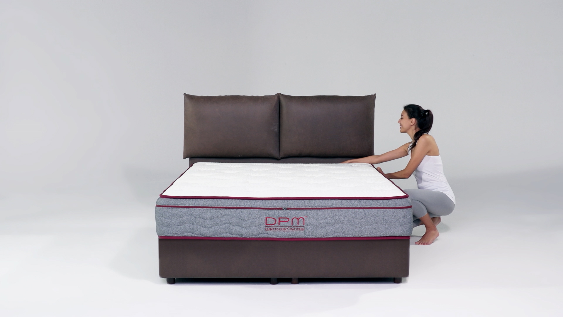 A woman in a white tank top and gray pants kneels beside a bed, pulling on the mattress in a minimalist room with a brown headboard and base, and light gray background.
