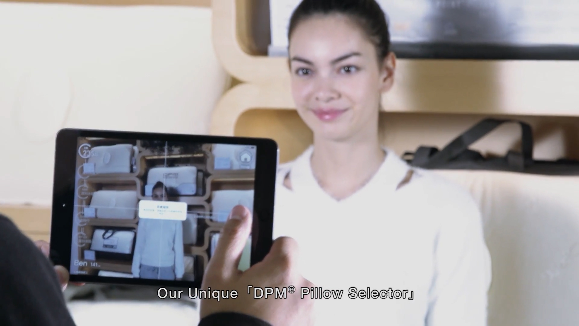 A person holds a tablet in front of a smiling woman, using an app to select a pillow. Shelves with pillows are visible in the background. On-screen text reads: "Our Unique 'DPM' Pillow Selector.