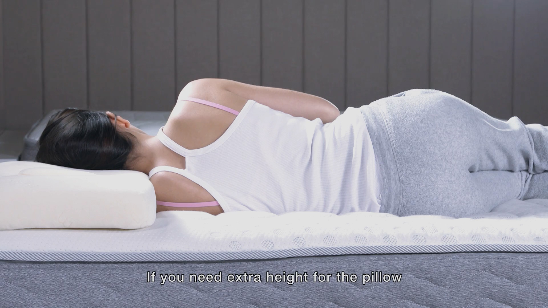 A woman in a white tank top and gray pants lies on her side on a mattress, resting her head on a white pillow. Text at the bottom reads, "If you need extra height for the pillow.