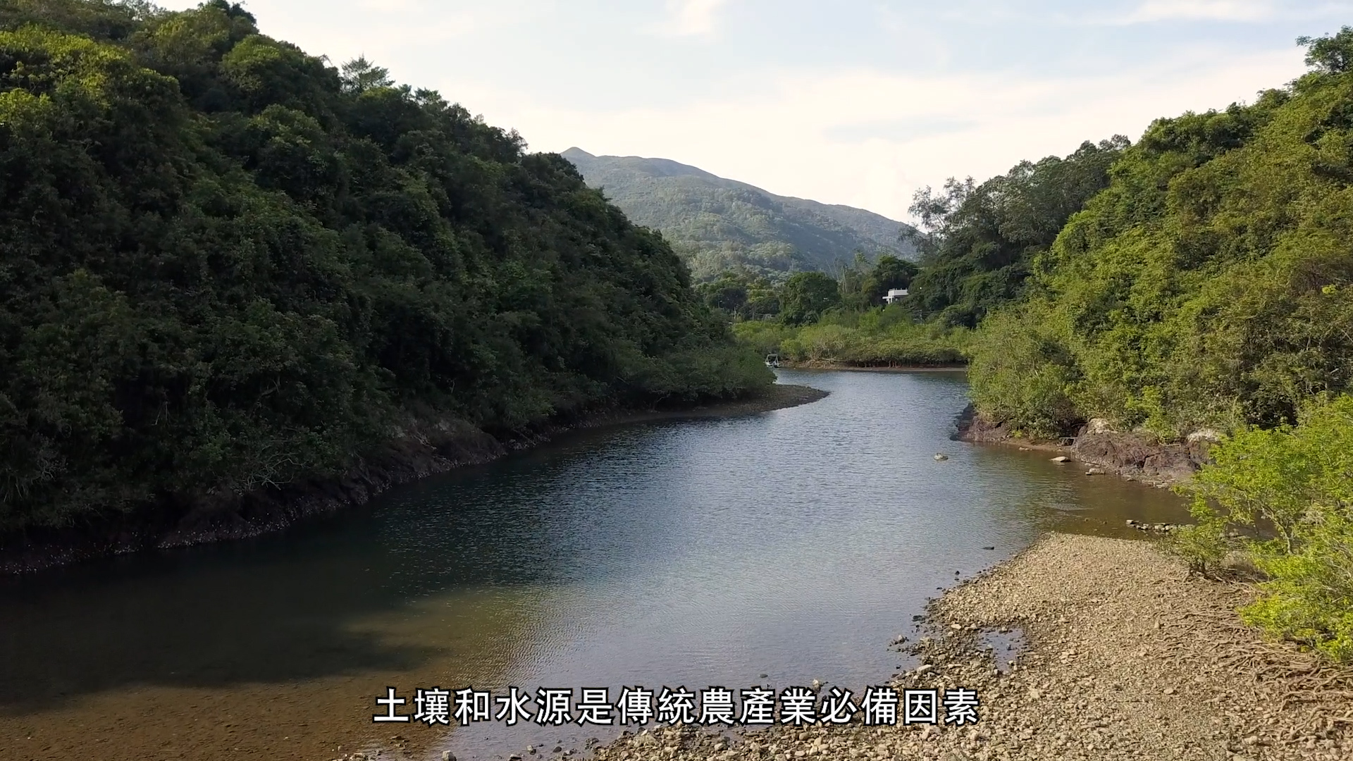 A peaceful river winds between green, tree-covered hills under a partly cloudy sky. Chinese subtitles appear at the bottom of the image.