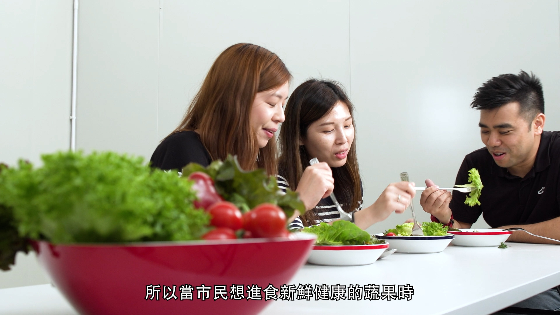 Three people sit at a white table eating fresh salad with forks. Bowls of vegetables, including lettuce and tomatoes, are on the table. Chinese subtitles are visible at the bottom of the image.