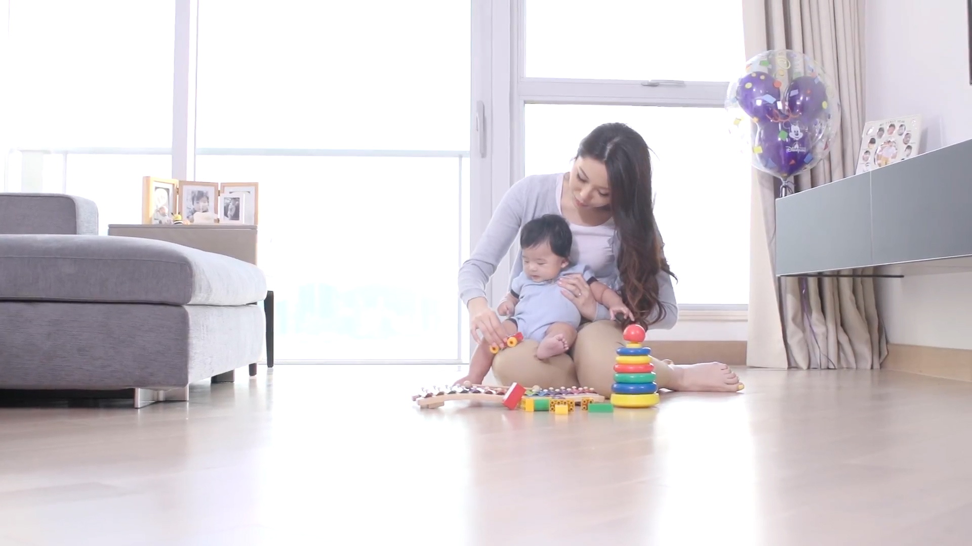 A woman sits on the floor with a baby on her lap, playing with colorful toys and blocks in a bright, sunlit living room.
