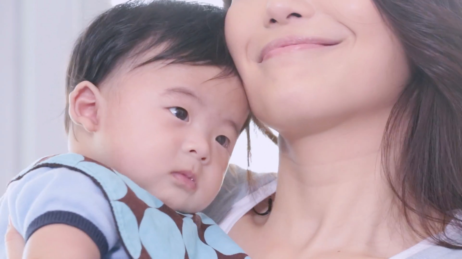 A woman smiles gently while holding a baby close to her cheek. The baby looks calm and relaxed, resting against the woman's shoulder. Both faces are partially visible, with a soft and warm atmosphere.