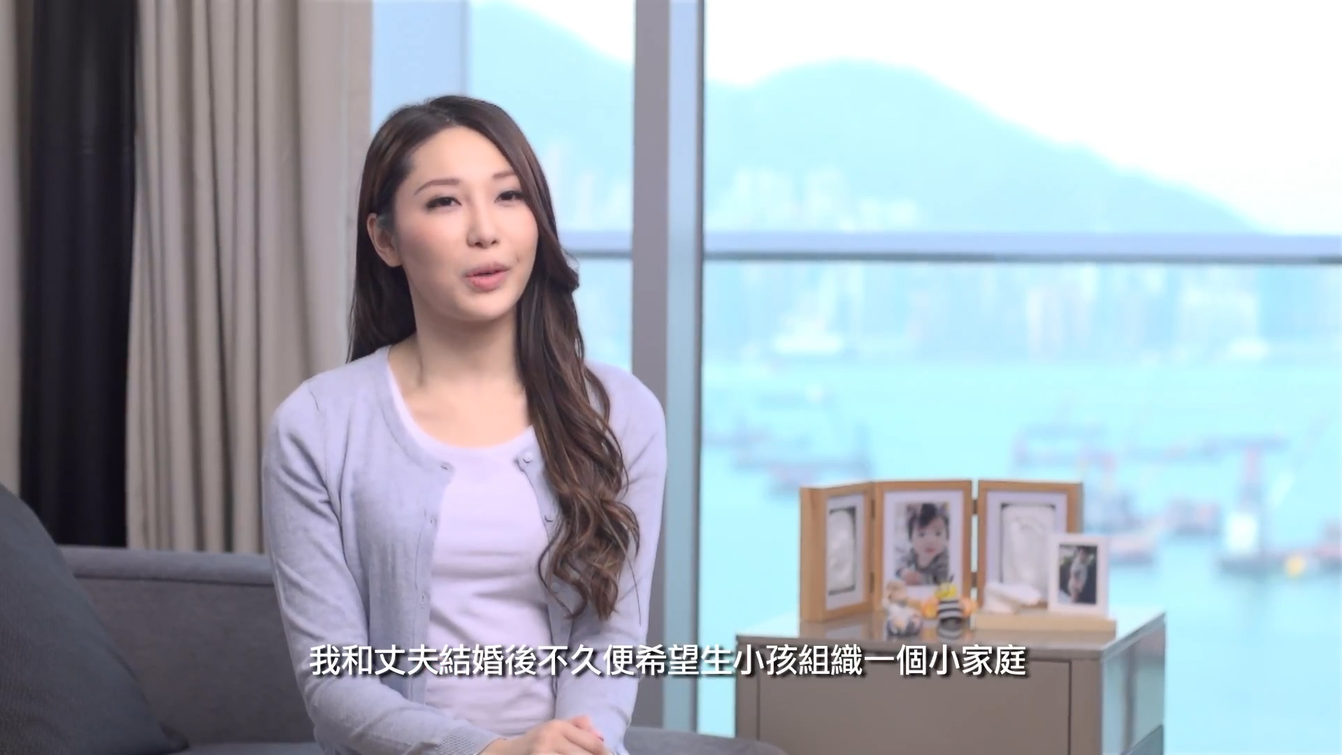 A woman with long hair sits indoors, speaking to the camera. Behind her are large windows showing a city and harbor view, as well as a table with several framed family photos. Chinese subtitles appear at the bottom of the image.