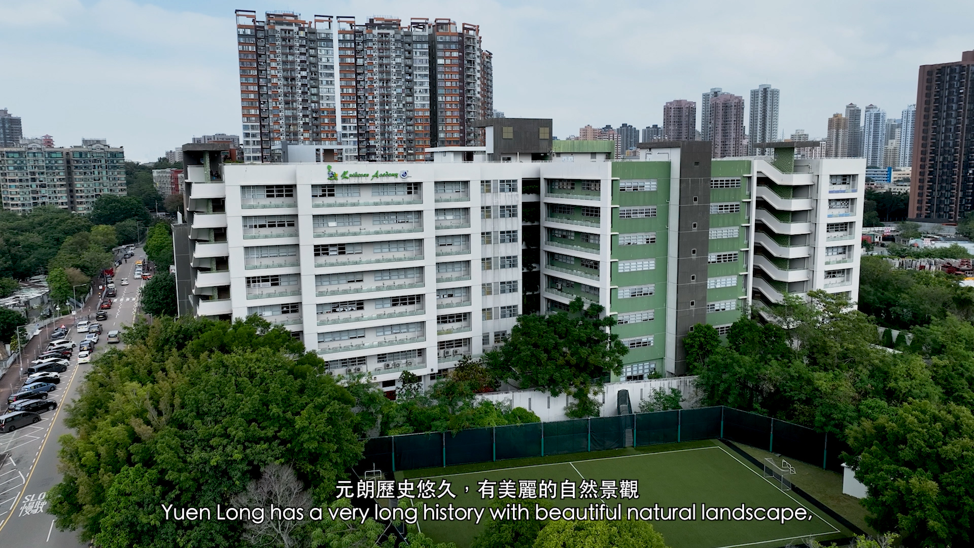 An aerial view of a multi-storey school building surrounded by trees, with a green sports field in front. High-rise apartments are visible in the background. Subtitles describe Yuen Long's long history and beautiful landscape.