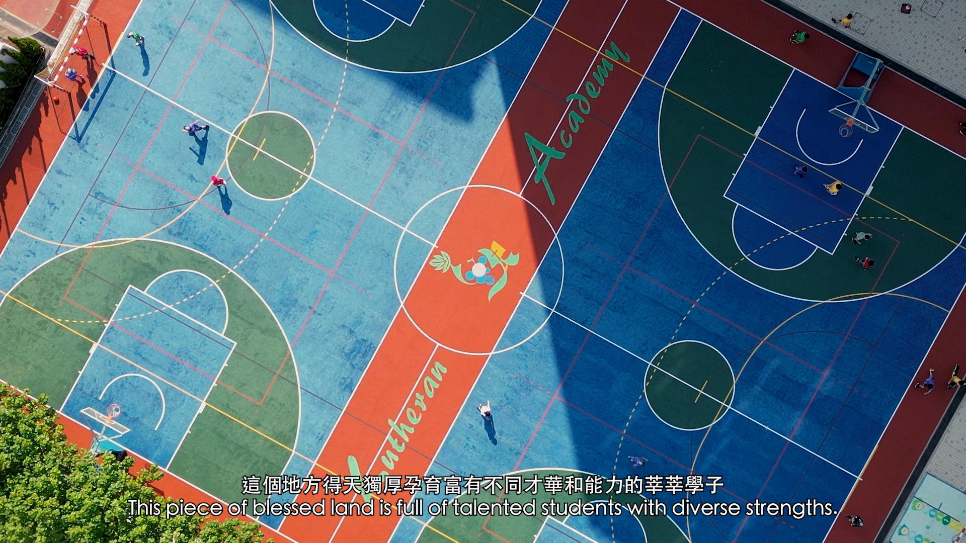 Aerial view of a colorful outdoor sports court with students playing; the court features the word "Academia," a rooster logo, and has both English and Chinese subtitles at the bottom.