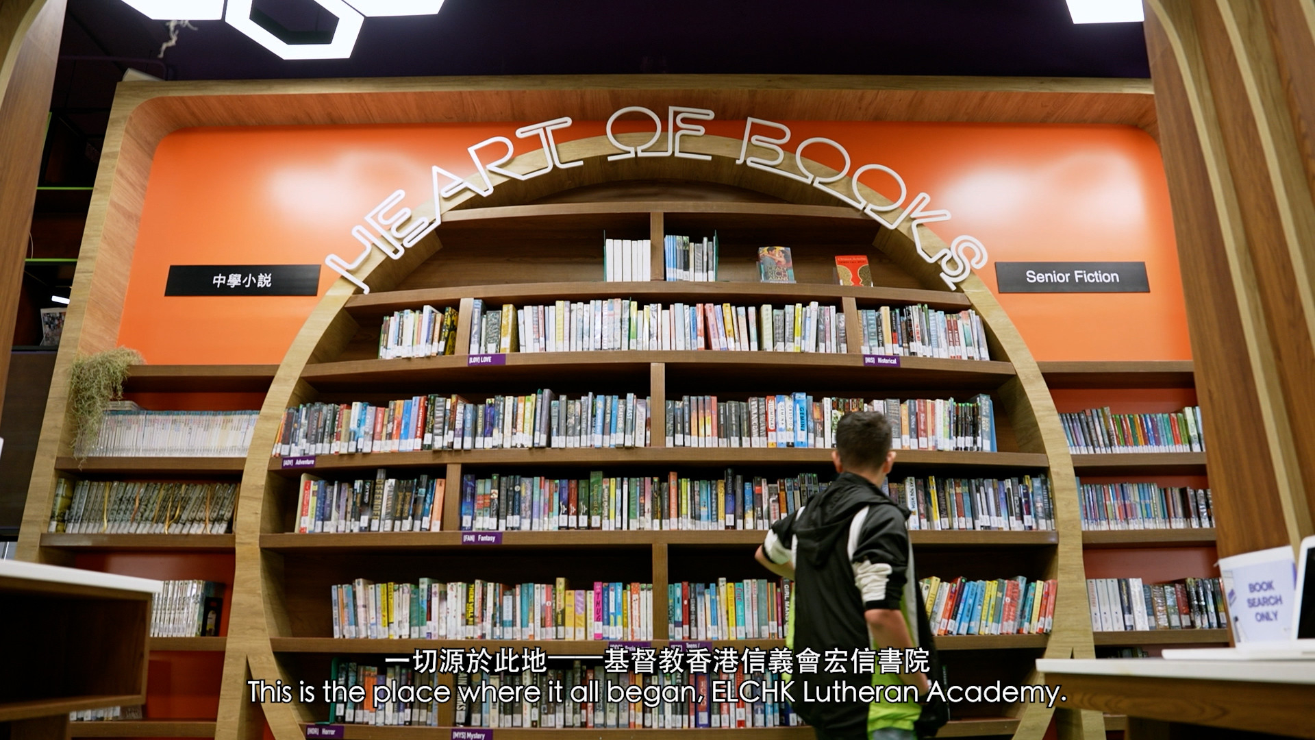 A child stands in front of a large bookshelf under an arch labeled "HEART OF BOOKS" in a library, reaching toward the books. Signs read "Senior Fiction" and "中文小說". Subtitles appear at the bottom of the image.