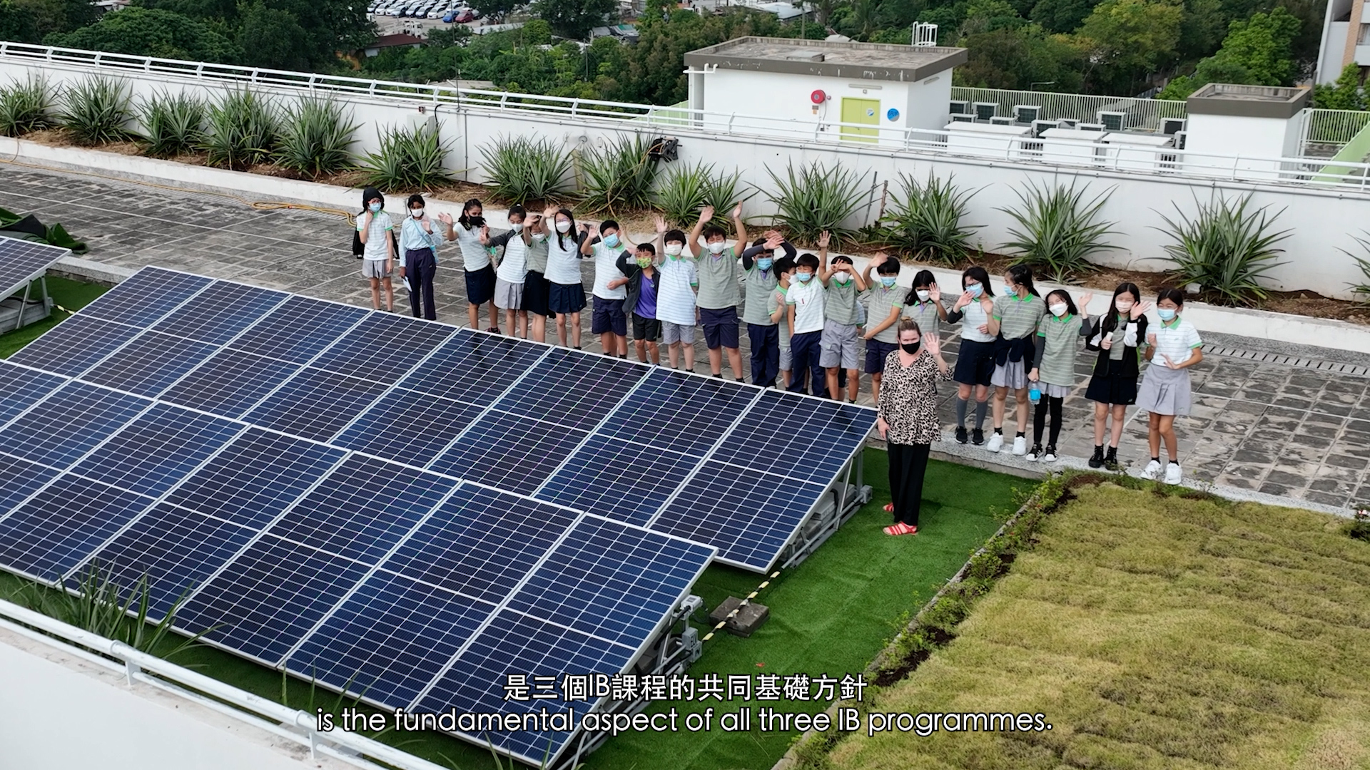 A group of students and a teacher stand near rooftop solar panels and greenery, waving at the camera. They are wearing masks and school uniforms. Subtitles at the bottom read: "is the fundamental aspect of all three IB programmes.