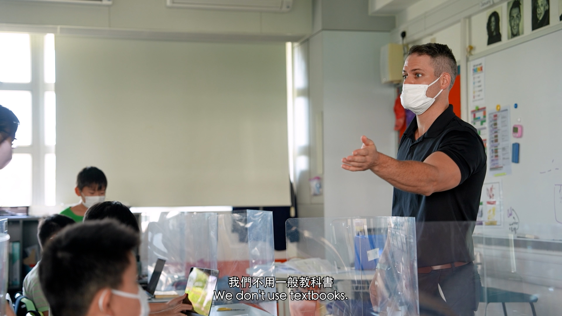 A teacher wearing a mask gestures while speaking to students in a classroom. The students, also masked, sit at desks with transparent dividers. Subtitles read: “We don’t use textbooks.”.
