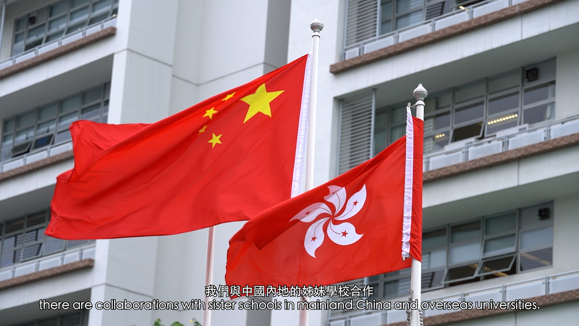 Two flags, the national flag of China and the flag of Hong Kong, are flying in front of a modern building. English and Chinese subtitles at the bottom mention collaborations with schools in mainland China and overseas.