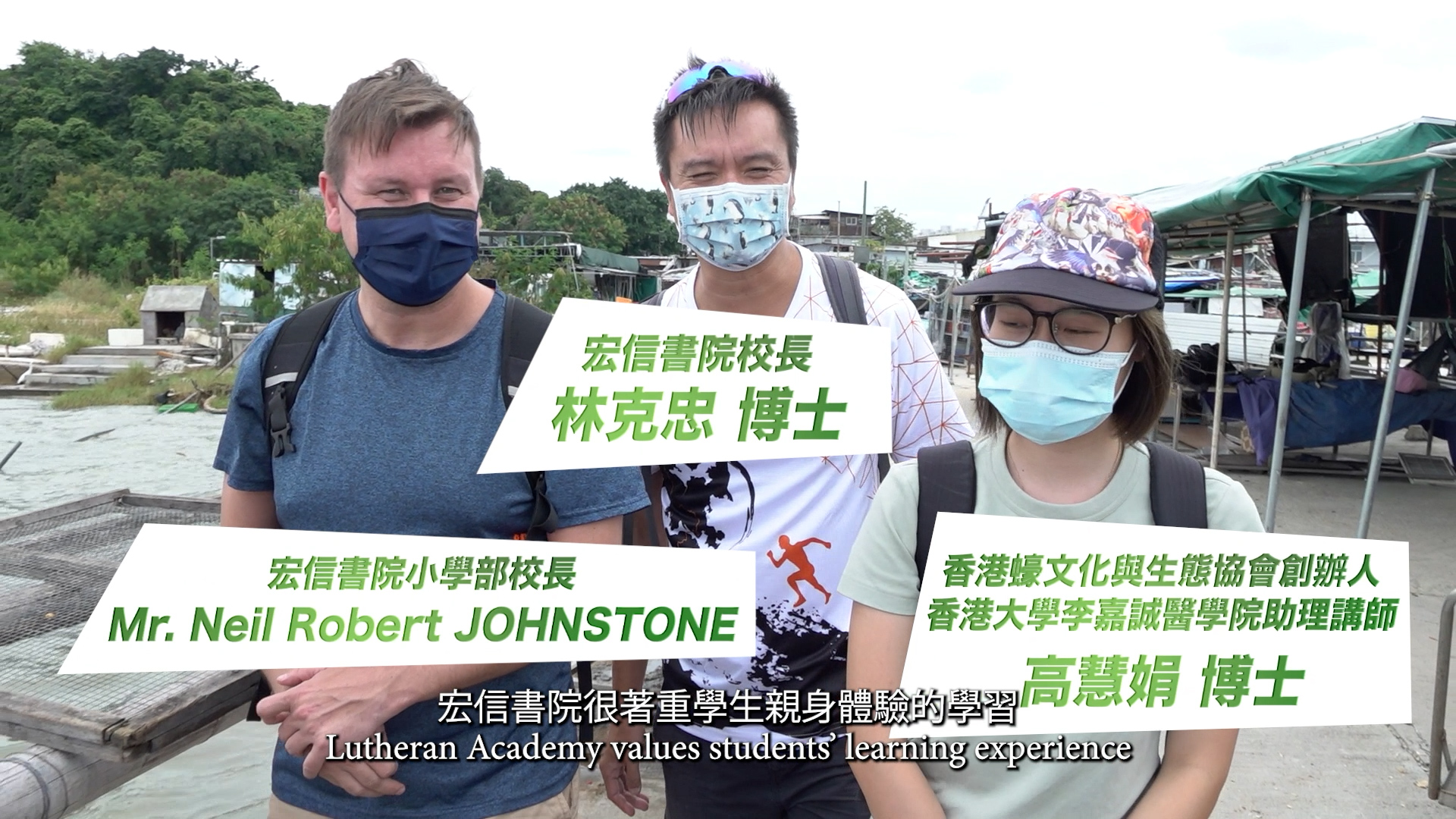 Three adults wearing masks stand outdoors near water with a hillside and boats in the background. On-screen text introduces them as Dr. Lin, Mr. Johnstone, and Dr. Fan. Subtitles mention hands-on learning at Lutheran Academy.