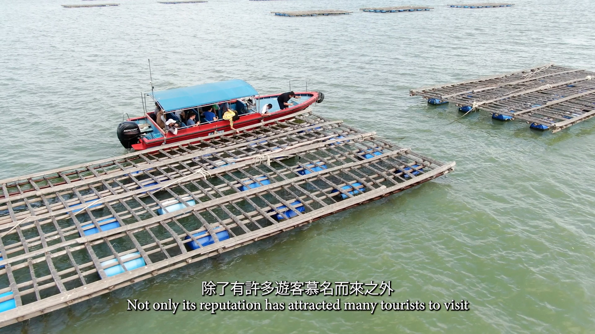A red boat with a blue canopy carries passengers past wooden fish farming platforms on green water. Subtitles in Chinese and English read, “Not only its reputation has attracted many tourists to visit.”.