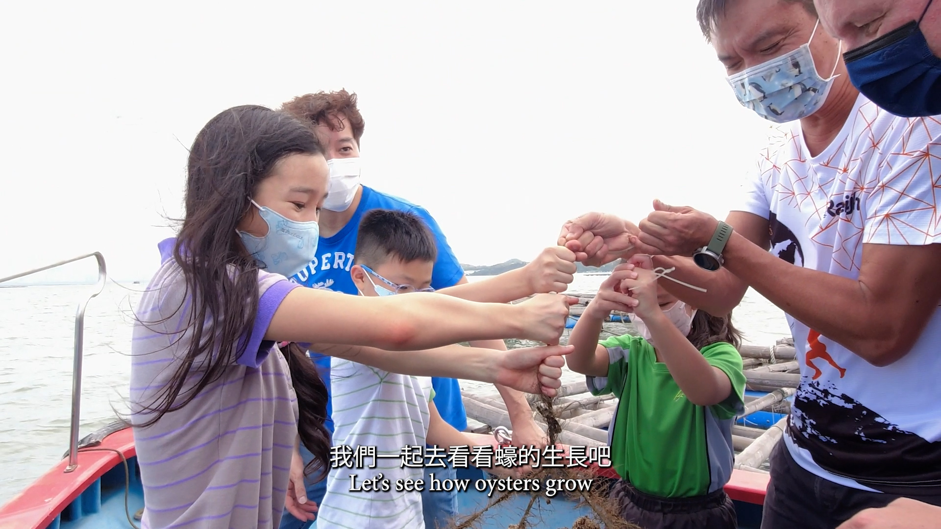 Four children and two adults, all wearing masks, examine oysters on a rope while standing on a boat. The sea and wooden poles are visible in the background. Subtitles read: "Let's see how oysters grow.