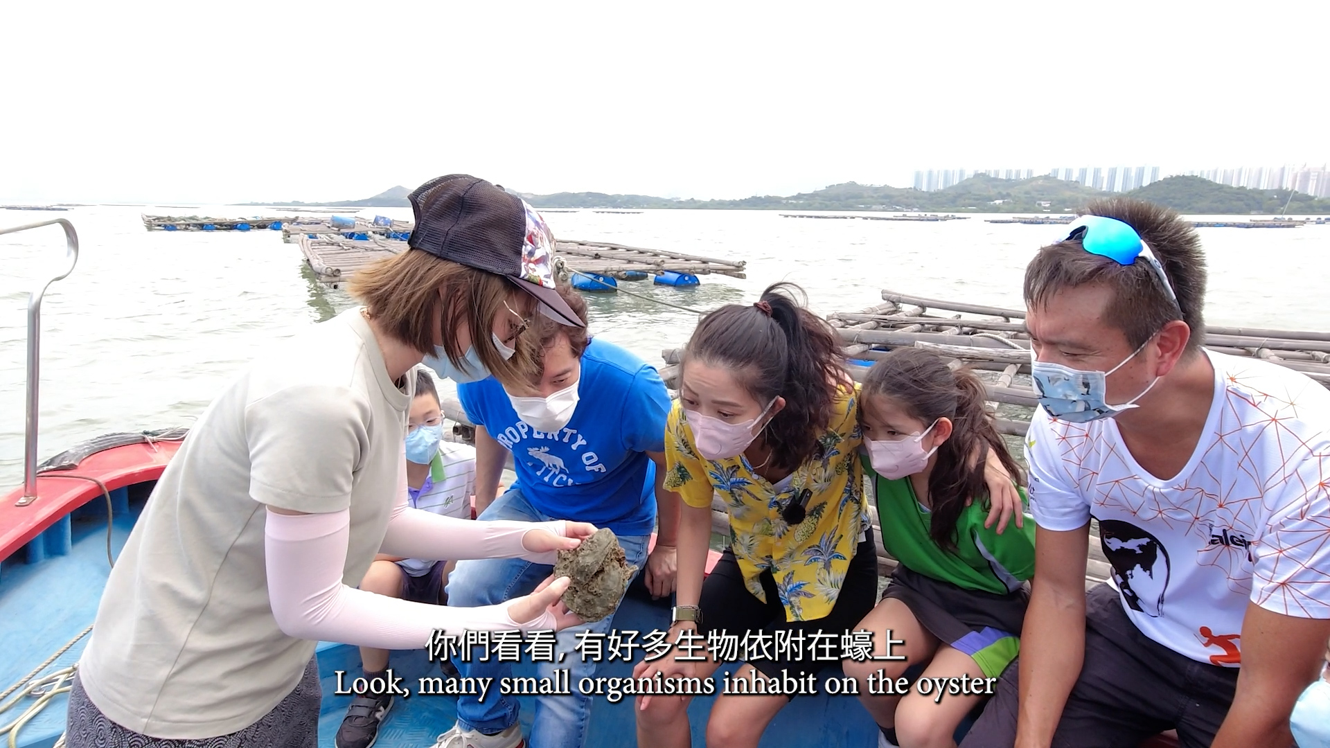 A woman on a boat shows an oyster to four children and one adult, all wearing face masks. Fish farms and city buildings are visible in the background. Subtitles read: "Look, many small organisms inhabit on the oyster.