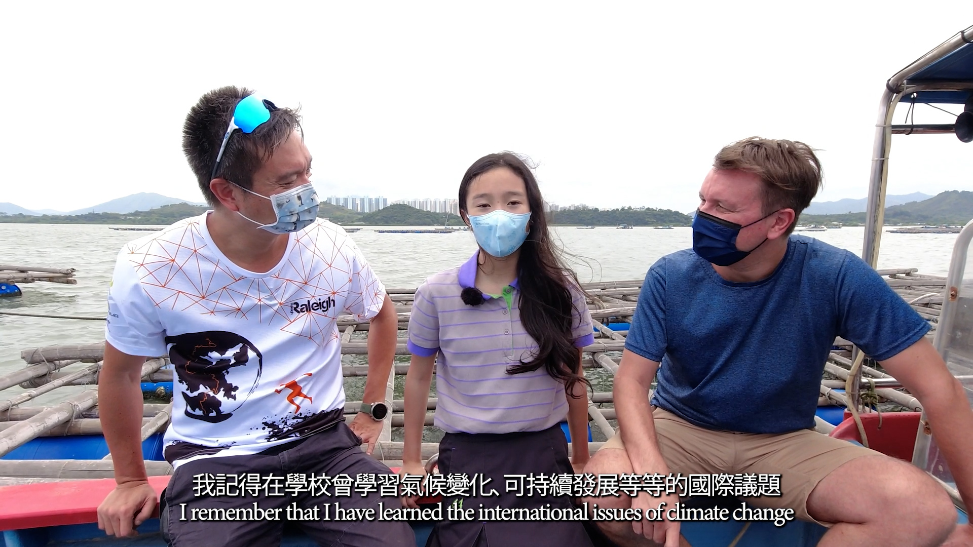 Three people wearing face masks sit on a boat with water and mountains in the background. The woman in the middle speaks, and subtitles about climate change and sustainability appear at the bottom.