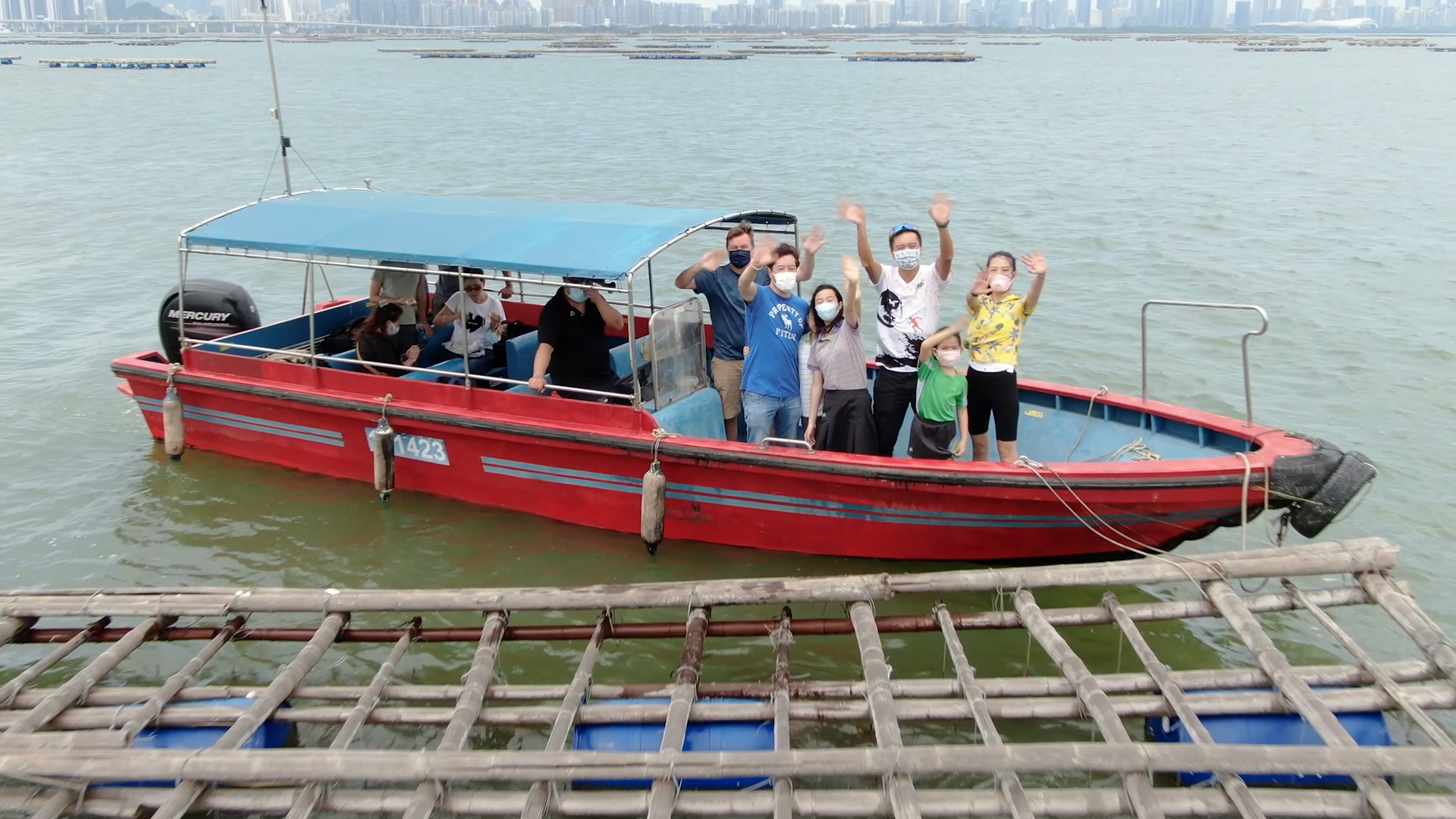 A group of people, some wearing masks, stand and wave on a red boat with a blue canopy on the water. The boat is near wooden structures, and a city skyline is visible in the distance across the water.