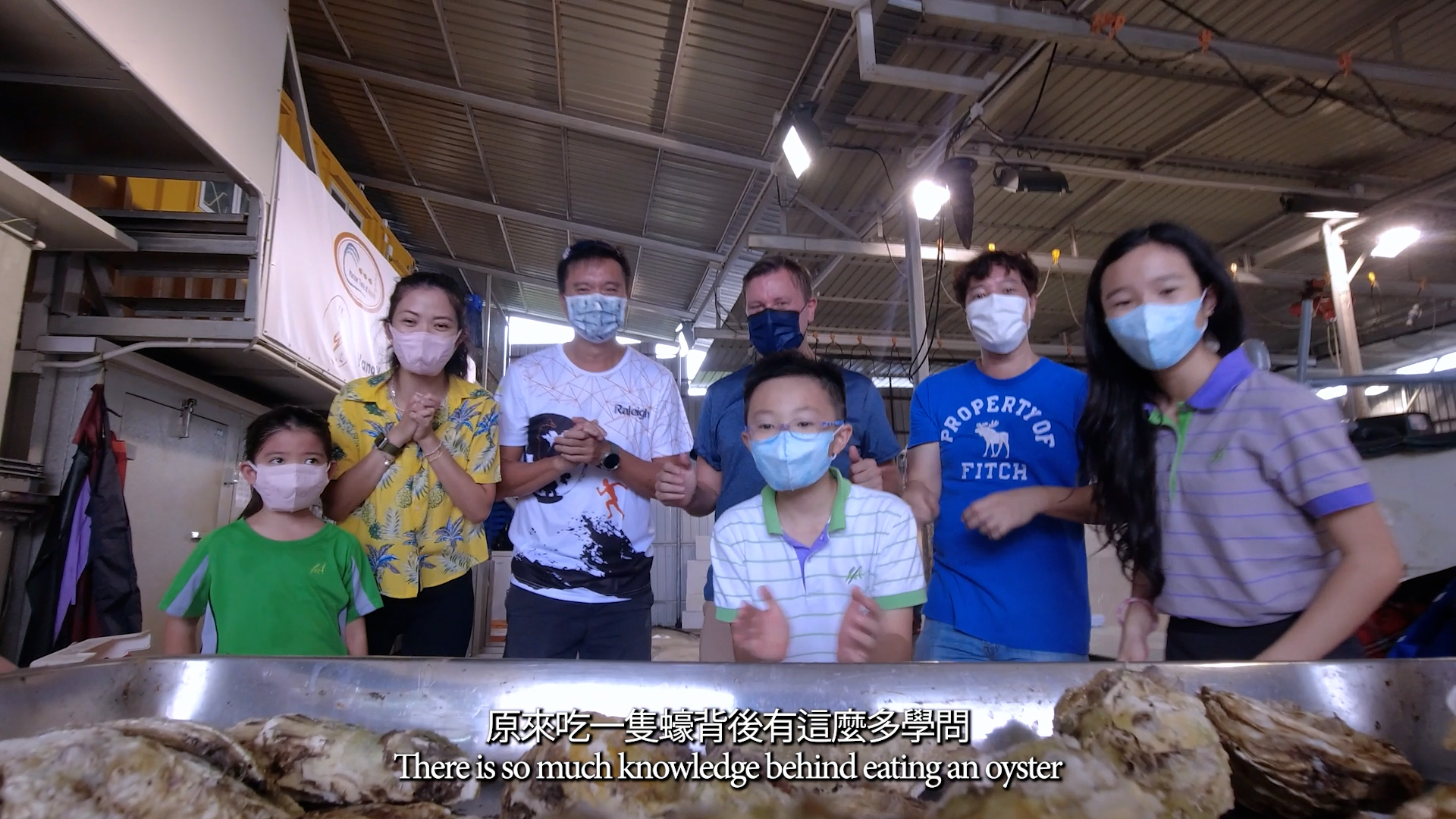 A group of seven people wearing masks stand indoors behind a table of oysters, smiling at the camera. Subtitles read: “There is so much knowledge behind eating an oyster.”.