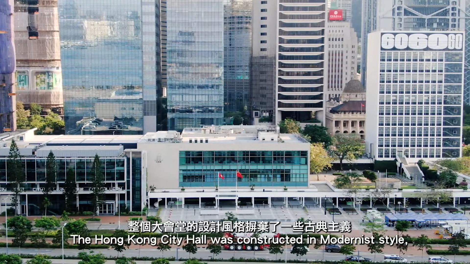 Aerial view of Hong Kong City Hall, a modernist building with glass windows and two flags, surrounded by tall skyscrapers. English and Chinese subtitles appear at the bottom, discussing the building’s architectural style.