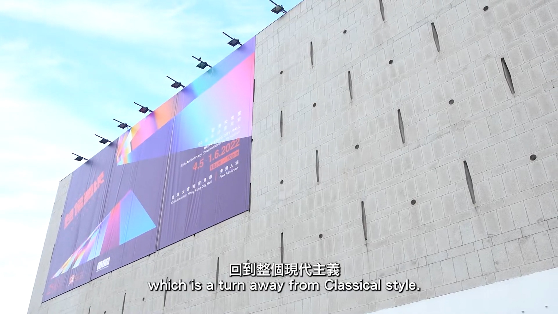 A modern concrete building with a large colorful banner promoting an event dated 4.5–12.2022. Subtitles at the bottom read: "which is a turn away from Classical style.