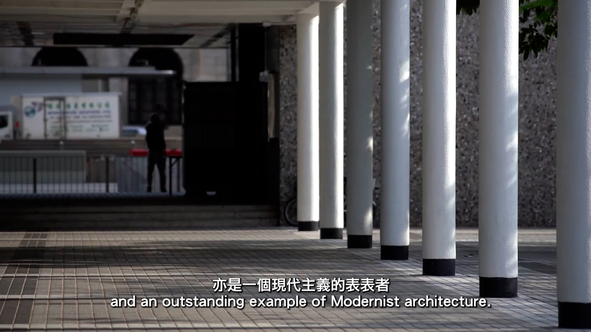 A row of white columns lines a covered walkway outside a modern building, with shadows on the tiled floor. Chinese and English subtitles read: "and an outstanding example of Modernist architecture.