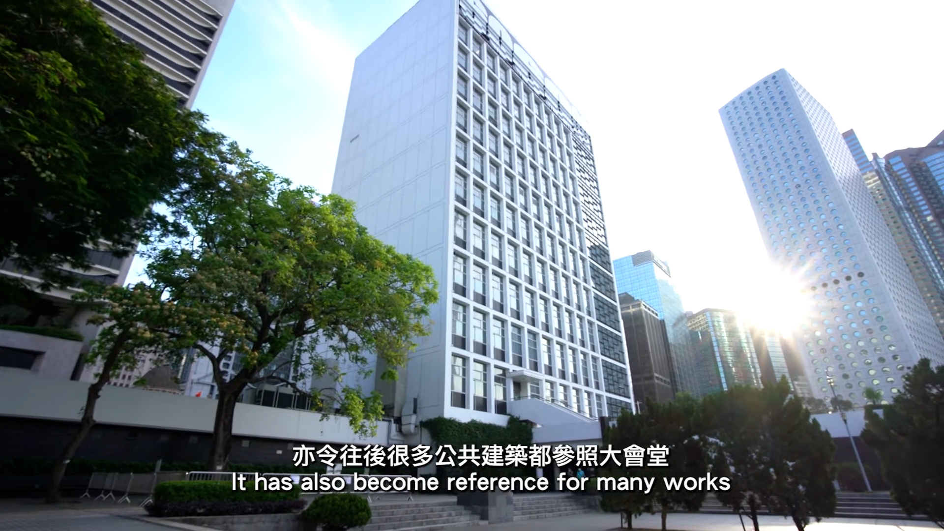 A modern white high-rise building stands among other skyscrapers and trees in an urban setting under a clear blue sky. Subtitles at the bottom read: “It has also become reference for many works.”.