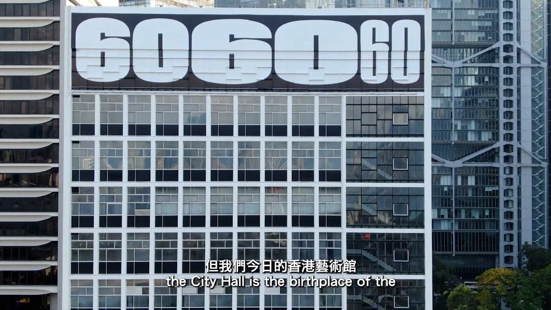 A modern building with large numbers "6060 60" displayed across the top floor. The façade features a grid of square windows. English and Chinese subtitles at the bottom read, "the City Hall is the birthplace of the".