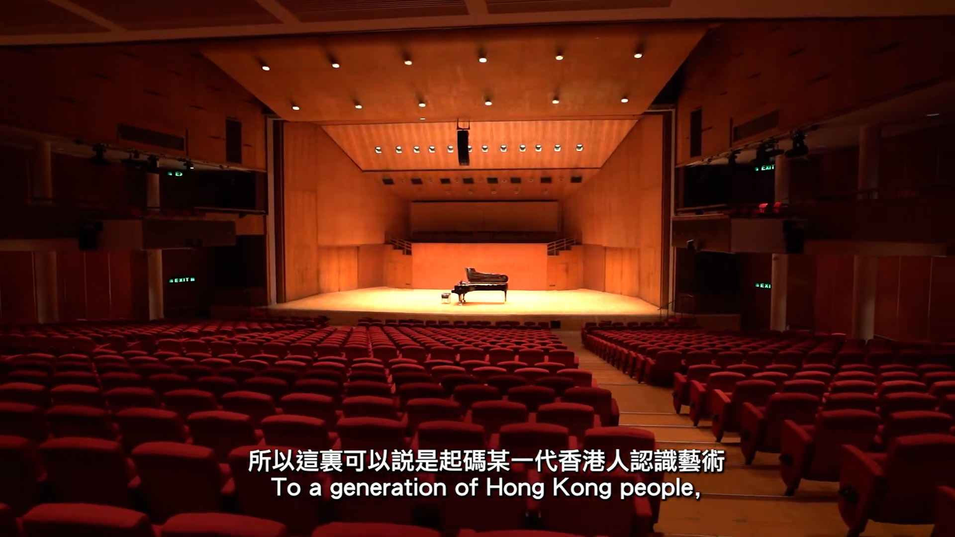 A grand piano sits on stage in an empty concert hall with red seats. Warm lighting illuminates the wooden interior. Chinese and English subtitles read: “To a generation of Hong Kong people,” along the bottom.