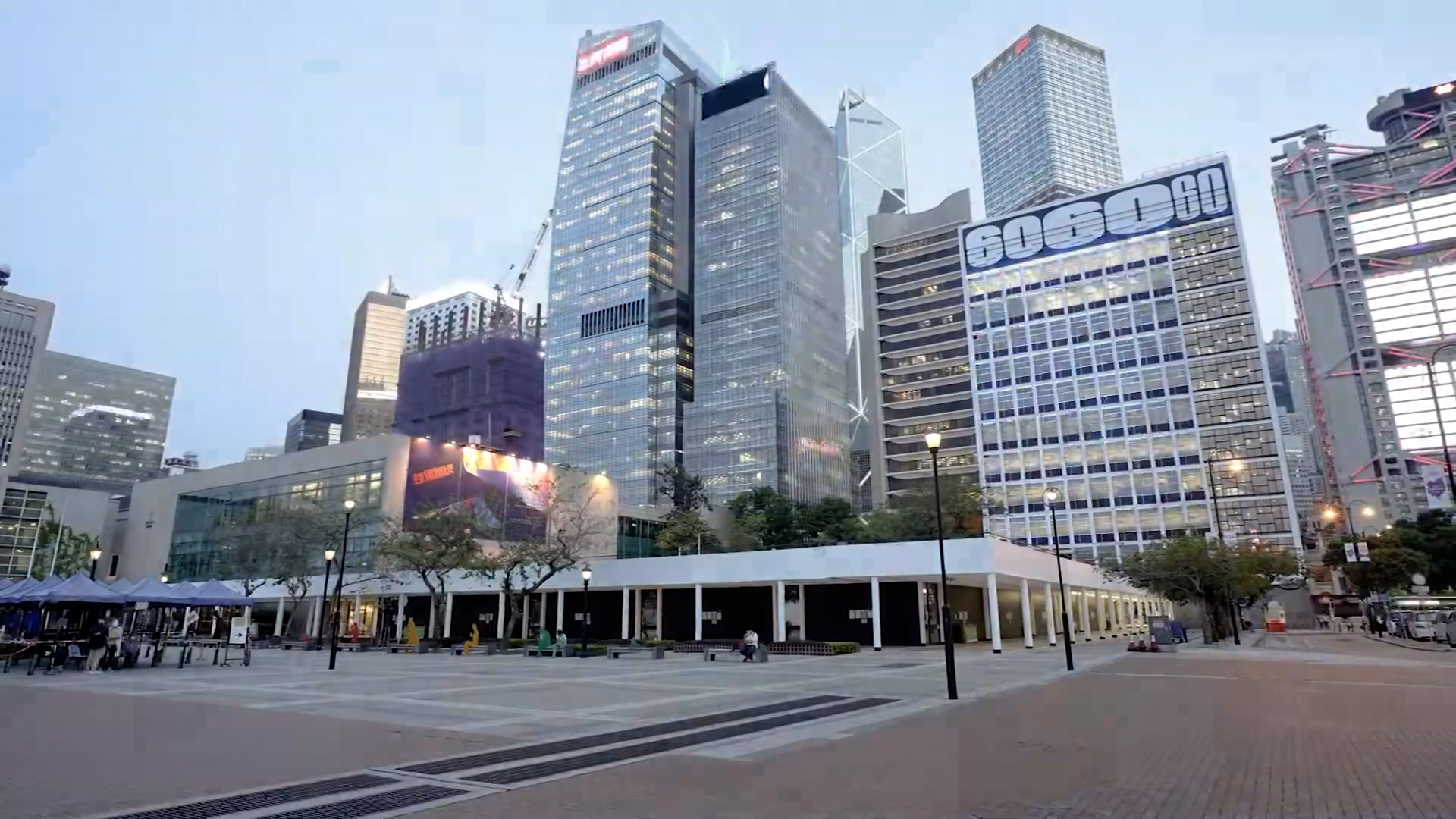 A city plaza with a few people walking, surrounded by modern high-rise buildings with glass facades and large advertisements, under a clear sky at dusk.