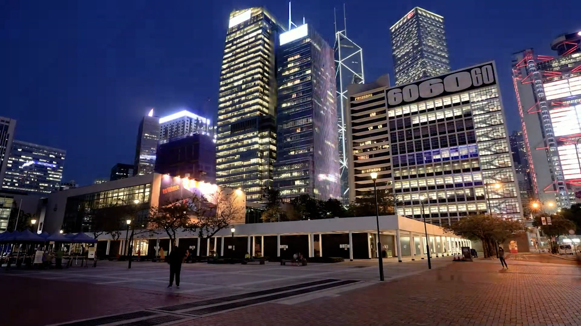 A cityscape at night featuring tall, illuminated skyscrapers, including a building marked "6060", with a few people walking on a spacious, paved plaza in the foreground.