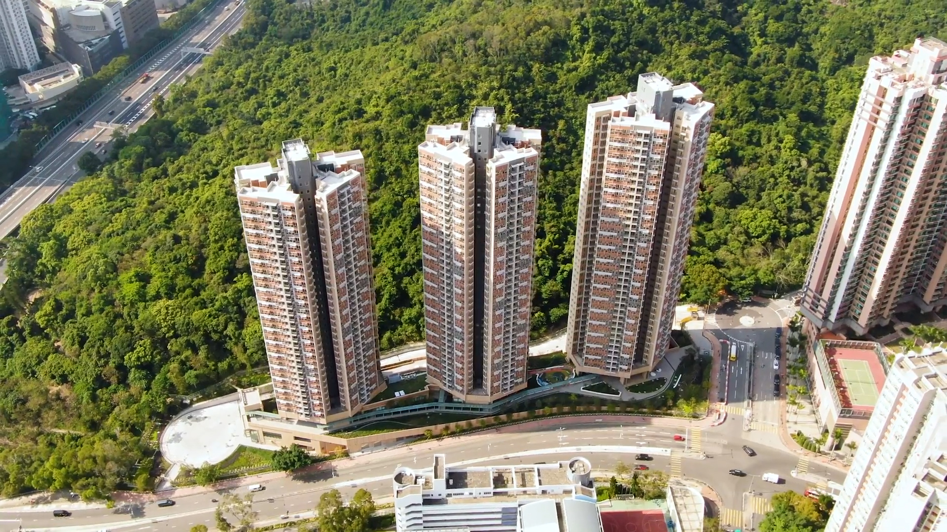 Aerial view of three tall residential apartment buildings standing in a row, surrounded by lush green forest and a curved road in an urban area.