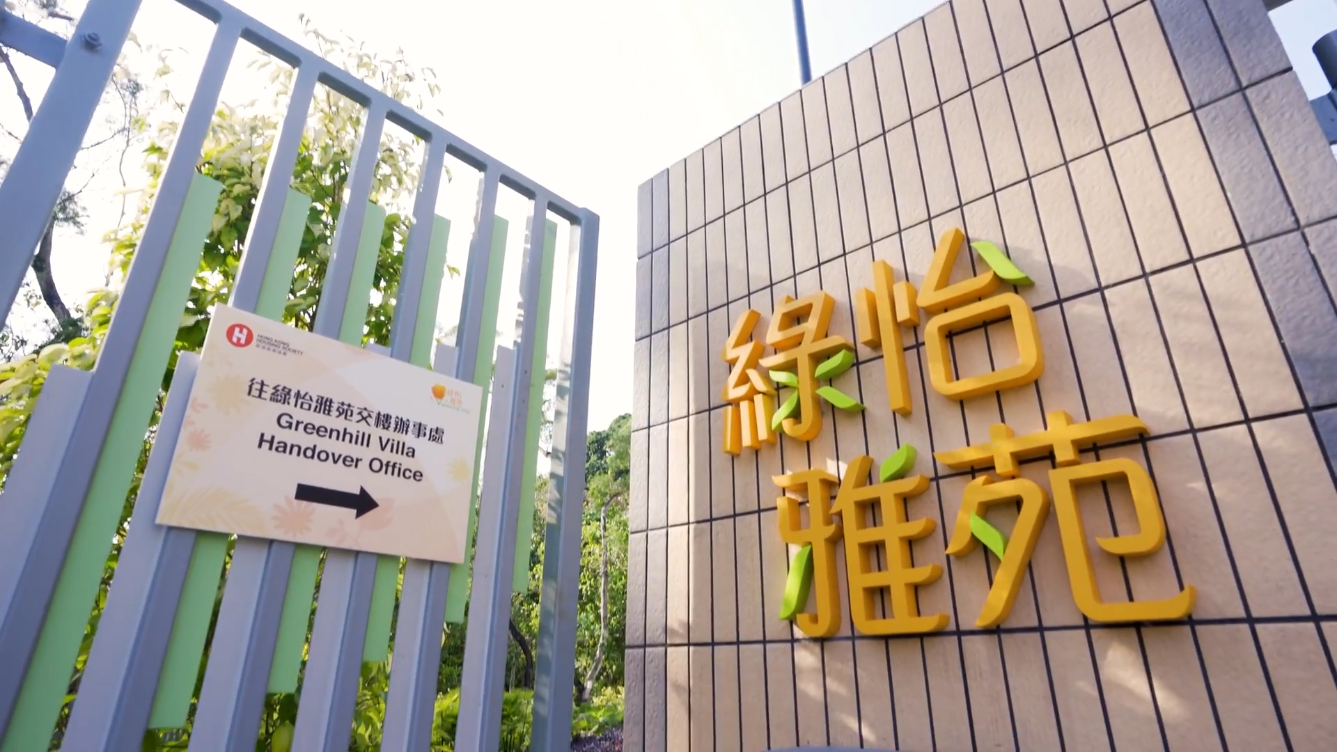 A gated entrance with a sign reading "Greenhill Villa Handover Office" and large yellow Chinese characters on a tiled wall, surrounded by greenery in bright sunlight.