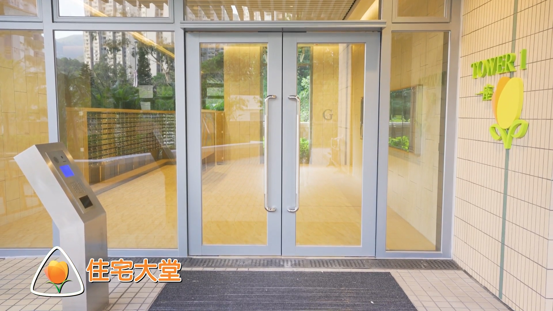 Glass entrance doors to a modern building with a gray doormat in front. There is a kiosk on the left and signage with Chinese text and “TOWER 1” on the right. Reflections and greenery are visible outside.